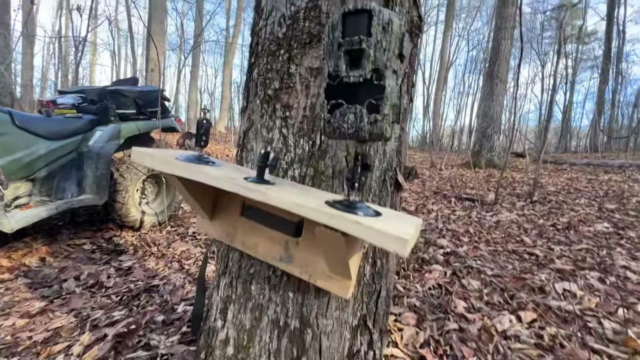 A Stealth Cam GMAX Vision trail camera on a tree in the woods, with an ATV parked beside it