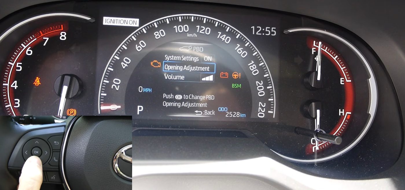 The 7-inch gauge screen of a 2019 RAV4 open to the PBD menu, showing System Settings ON, Opening Adjustment and Volume, with an inset of a thumb on the OK button of the steering wheel arrow pad
