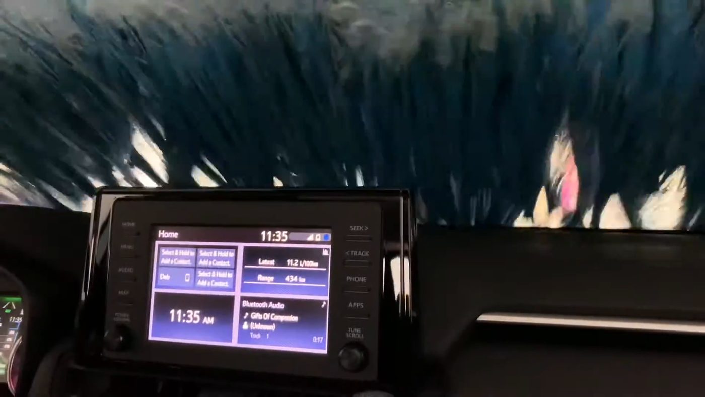 View from the driver's seat of a 2020 RAV4 on an automatic conveyor car wash, with a spinning brush covering the windshield above the RAV4's touch screen