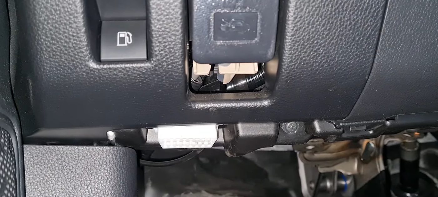 The lower dash of a 2020 RAV4 Hybrid on the driver's side, with the fuel door button and the hood release lever above and Toyota's white OBD-II plug clipped into the bottom edge of the dash
