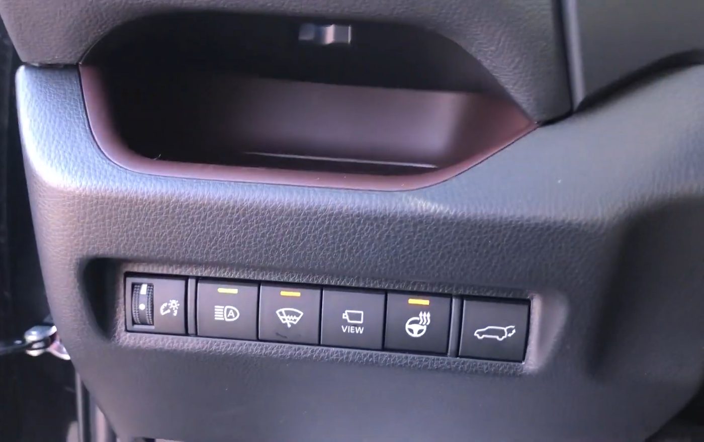 The row of buttons low on the dash left of the steering column in a 2020 RAV4 Limited, from left to right the dash brightness dial, Automatic High Beam, wiper de-icer, VIEW camera, heated steering wheel and power liftgate buttons, with the de-icer and heated steering wheel lights on