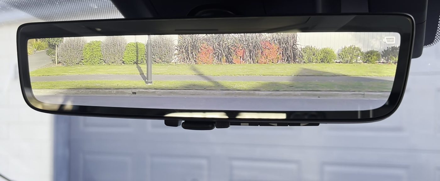 The digital rearview mirror on a 2023 to 2025 RAV4 with the camera picture on, the small camera icon in the top right corner of the screen, and the lever and buttons along the bottom edge of the mirror