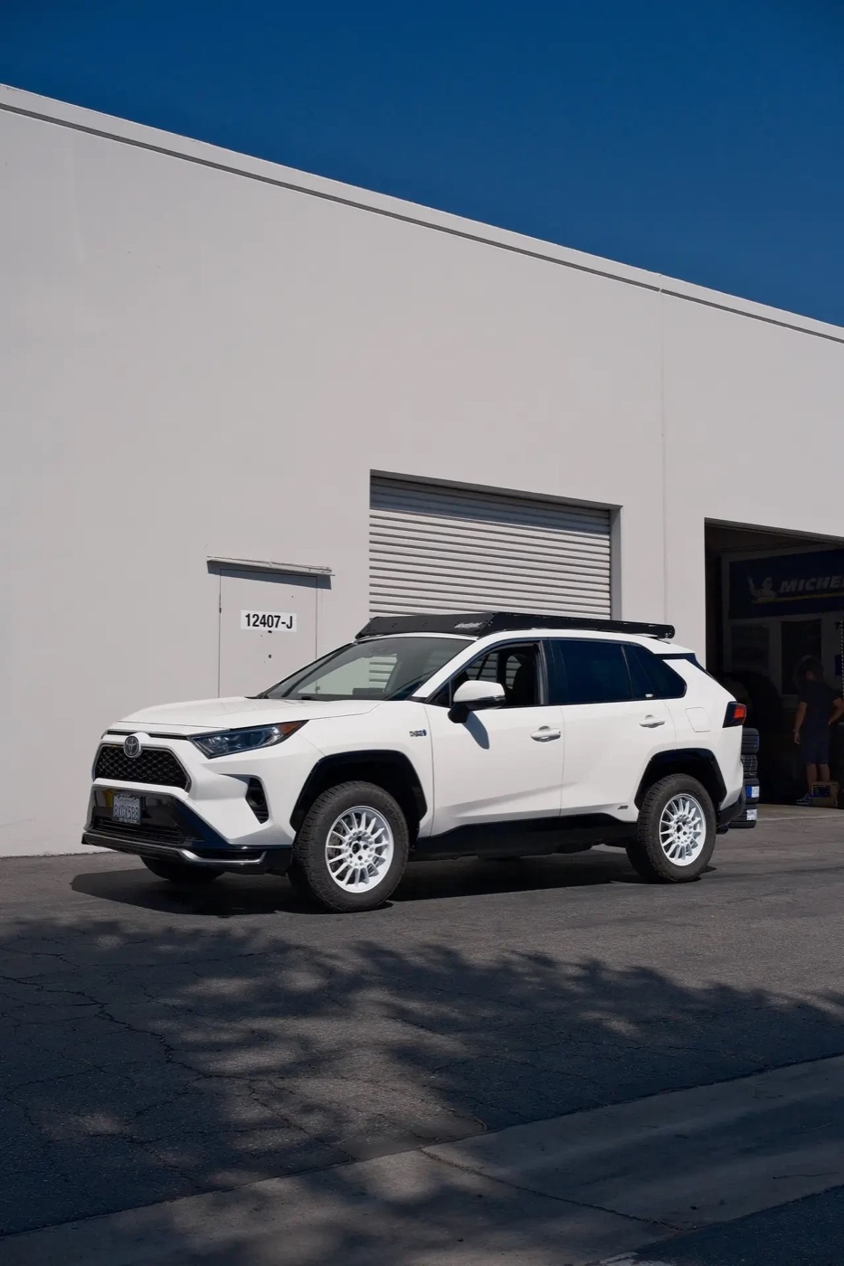 A white RAV4 Prime on white multi-spoke wheels and all-terrain tires with a low roof rack
